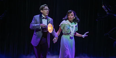 A man in a plaid suit holds a flashlight and a woman’s hand. The woman wears a light green dress. Both appear startled on a dark stage with dramatic lighting.