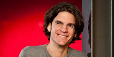 A person with medium-length brown hair wearing a gray sweater is smiling, standing in front of a red background and next to a dark wall.