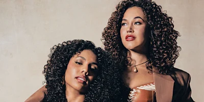 Two women with curly hair pose together against a neutral background; one has her head resting on the other's shoulder, both looking off camera.