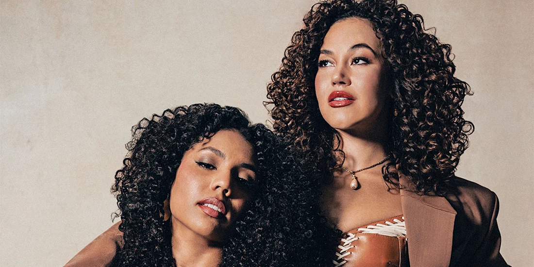 Two women with curly hair pose together against a neutral background; one has her head resting on the other's shoulder, both looking off camera.