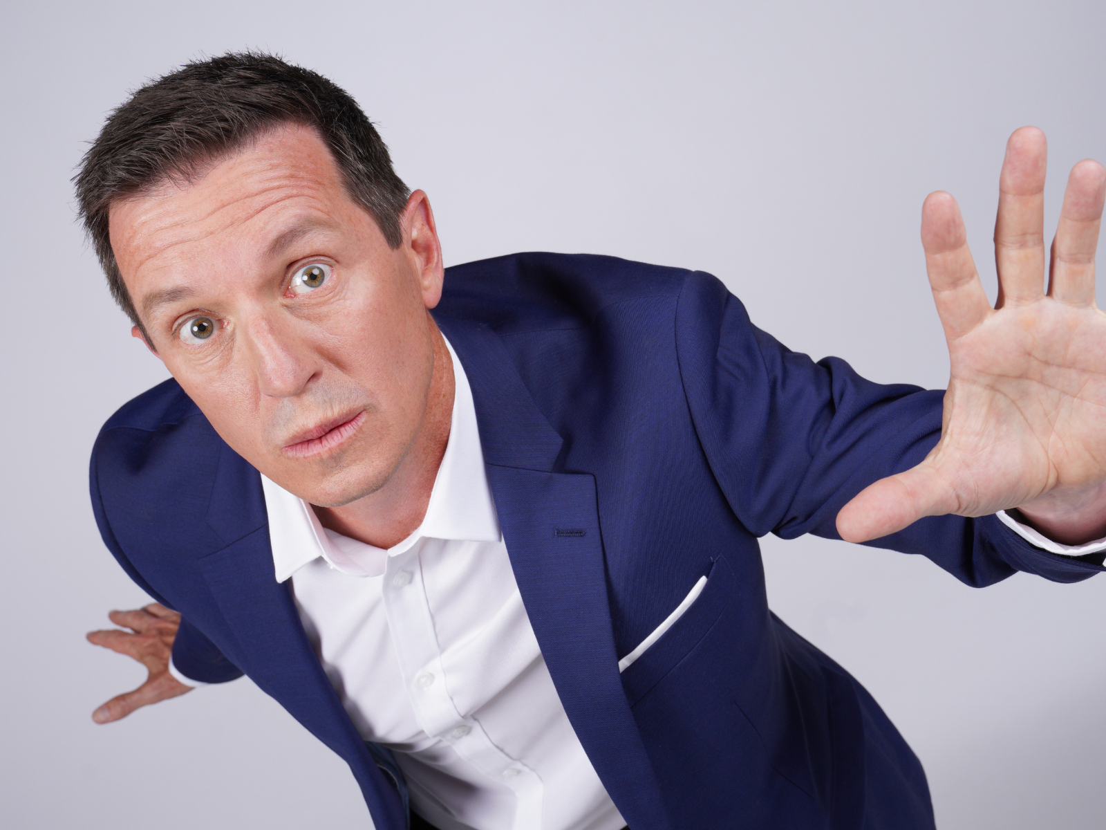 A man in a blue suit and white shirt leans forward with a serious expression, holding one hand up in front of him as if signaling to stop.