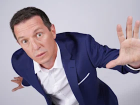 A man in a blue suit and white shirt leans forward with a serious expression, holding one hand up in front of him as if signaling to stop.