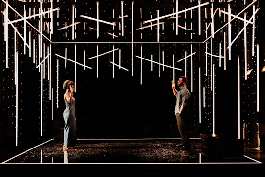 Two people stand facing each other on a stage with hanging vertical lights and geometric lighting above and below, creating a modern, abstract atmosphere.