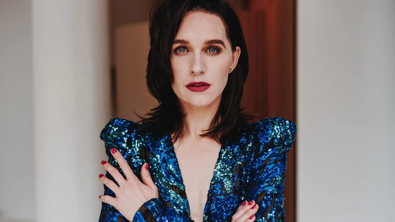 Woman with straight dark hair and red lipstick wearing a blue sequin dress, arms crossed over chest, standing indoors against a neutral background.