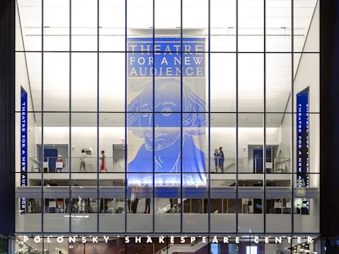 Glass facade of Polonsky Shakespeare Center at night, with large blue banner displaying Shakespeare's face and "Theatre for a New Audience" hanging inside the building.