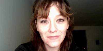A woman with shoulder-length brown hair and bangs looks directly at the camera, smiling slightly, in a room with neutral-colored walls.