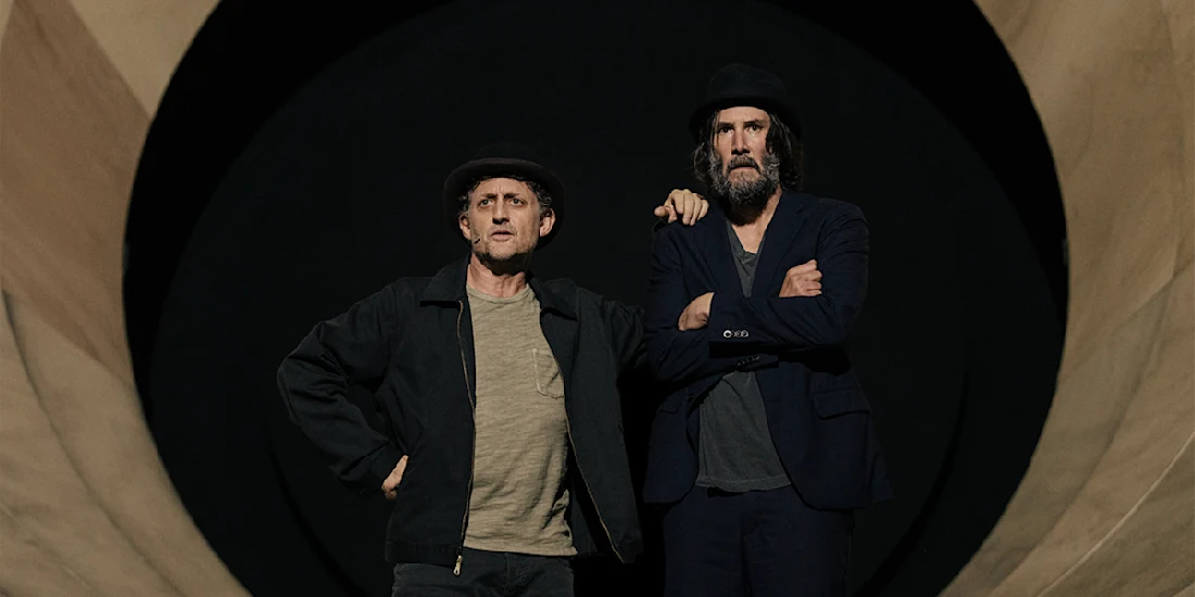 Two men stand side by side with serious expressions, both wearing dark jackets and hats, in front of a large circular backdrop.