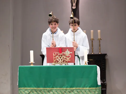 Two altar servers in white robes stand behind a green-covered altar, each holding a brass candlestick. Candles and a red book are on the altar, with a crucifix and candles in the background.