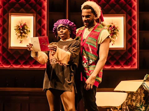 Two people stand together indoors; one reads a card and wears a purple bonnet, oversized shirt, and shorts, while the other wears a festive sleeveless sweater and Santa hat. Holiday decorations are visible.