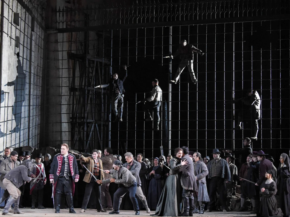 Production photo of Il Trovatore in New York, showing a dramatic stage scene depicting actors in period costumes, including soldiers and civilians. Some actors are suspended against a large metal grid backdrop, creating a tense atmosphere.