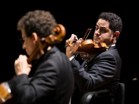 Two musicians in black suits play string instruments; one plays a cello in the foreground, and the other plays a violin in the background.