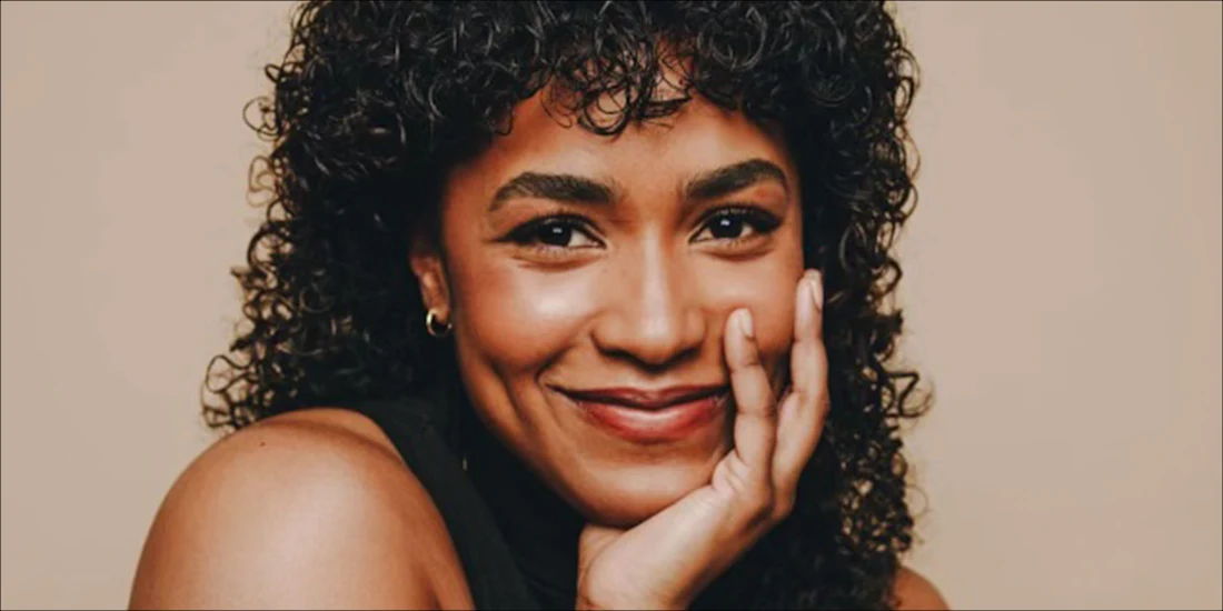 A person with curly dark hair and glowing skin smiles with their hand resting on their cheek against a plain beige background.