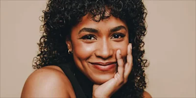 A person with curly dark hair and glowing skin smiles with their hand resting on their cheek against a plain beige background.