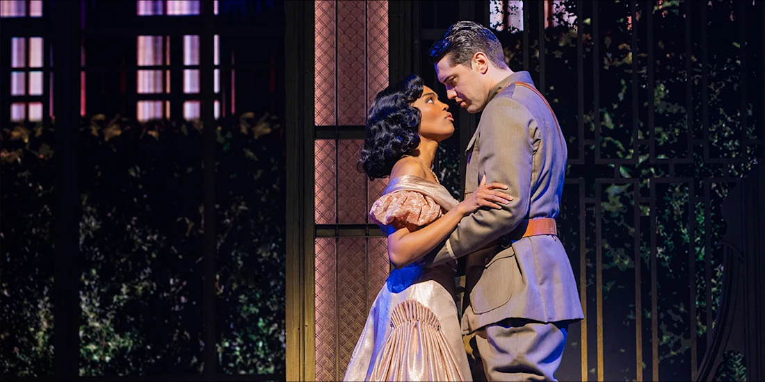 A woman in a pink dress and a man in a gray military uniform stand close together onstage, looking into each other’s eyes, with a decorative gate and vines in the background.
