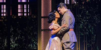 A woman in a pink dress and a man in a gray military uniform stand close together onstage, looking into each other’s eyes, with a decorative gate and vines in the background.