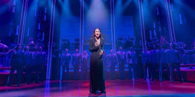 A woman stands and sings at center stage in dramatic blue and purple lighting, with a chorus of people in suits standing behind her on a theater set.