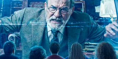 Four people sit facing a large screen displaying a serious older man with glasses and a beard, wearing a suit and tie, gesturing intensely.