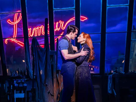 A man and a woman stand close together, gazing into each other's eyes on a stage with a neon "L'amour" sign and blue-lit windows in the background.
