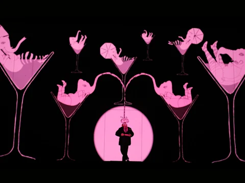 A person stands on stage in front of a pink-lit backdrop featuring illustrated figures lounging inside oversized cocktail glasses.