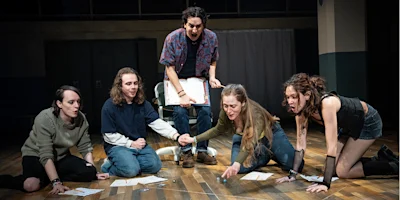 Five people on a stage sit or kneel in a semi-circle on a wooden floor, holding character sheets and rolling dice, suggesting a tabletop role-playing game scene.