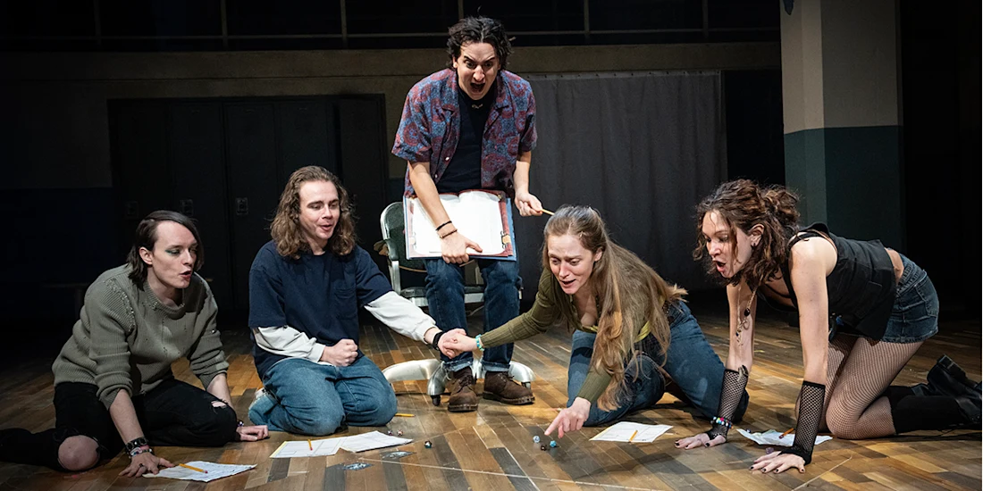 Five people on a stage sit or kneel in a semi-circle on a wooden floor, holding character sheets and rolling dice, suggesting a tabletop role-playing game scene.