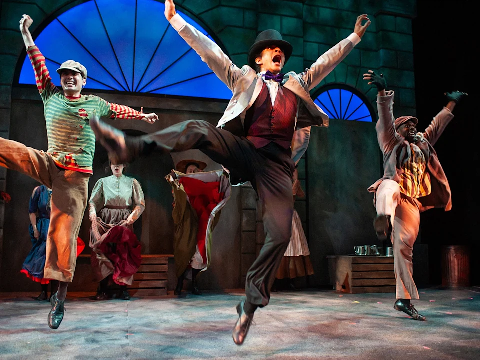 A group of performers in colorful costumes leap mid-air during a lively stage musical, set against a backdrop with arched windows.