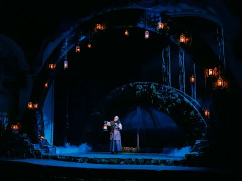 A person in costume stands on a dimly lit theater stage holding a lantern, surrounded by hanging lanterns and dramatic arches.