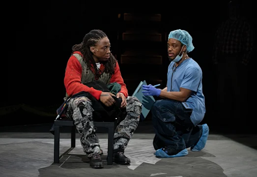 Two people are on stage; one sits wearing a red sweater and patterned pants, while the other kneels in blue medical scrubs, gloves, and a hair cap, holding a clipboard.