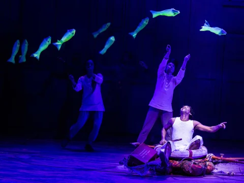 Three performers gesture towards glowing fish suspended in the air, while a fourth person sits on the floor surrounded by props. 