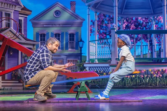 An adult and a child sit on opposite ends of a seesaw on a stage set resembling a colorful suburban neighborhood with houses and a gazebo.