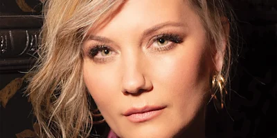 A close-up of a person with light hair, subtle makeup, and gold hoop earrings, looking directly at the camera against a dark background.