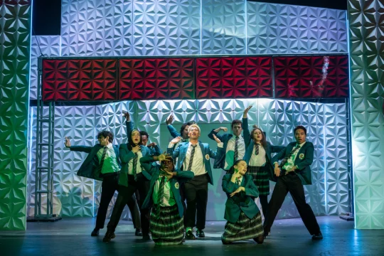 A group of performers in green blazers and plaid skirts or pants are on stage, striking energetic poses against a geometric backdrop with red and white sections.