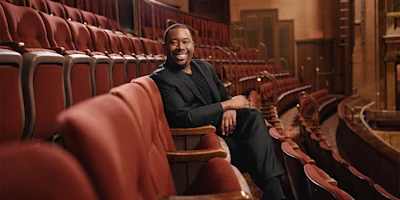 A man in a black suit sits smiling in an empty theater, surrounded by rows of red seats.