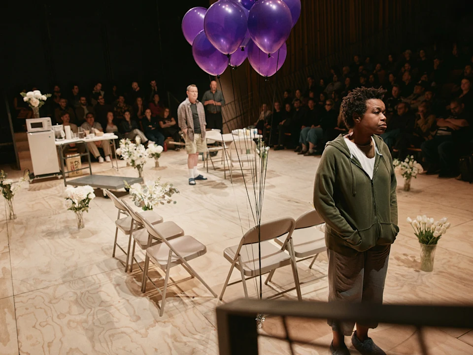 A woman stands near folding chairs and purple balloons on a stage set with flowers, while a man and an audience are visible in the background.