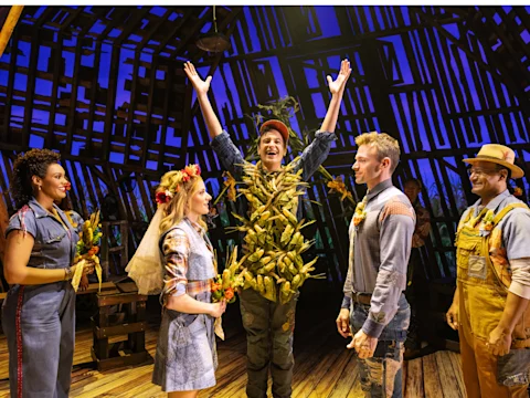 Five actors perform on stage; one actor dressed in a corn-themed costume raises their arms as others, in rustic attire, look on and hold small bouquets under dramatic lighting.