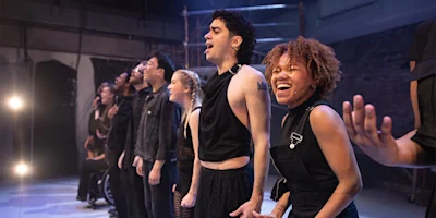 A diverse group of performers in black clothing stand in a line on stage, singing or speaking passionately under bright stage lights.