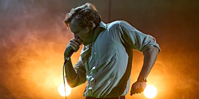 A man in a button-down shirt sings into a microphone on stage, with bright yellow-orange lights and smoke in the background.