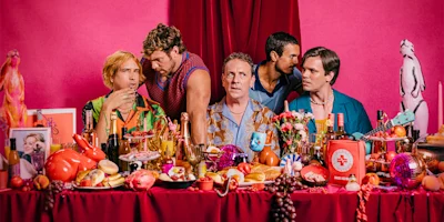 Five men sit at a table covered with assorted food, drinks, and colorful objects against a pink background with red curtains and a penguin statue in the corner.