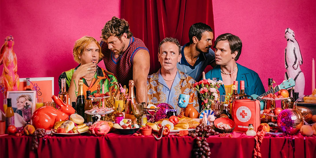 Five men sit at a table covered with assorted food, drinks, and colorful objects against a pink background with red curtains and a penguin statue in the corner.