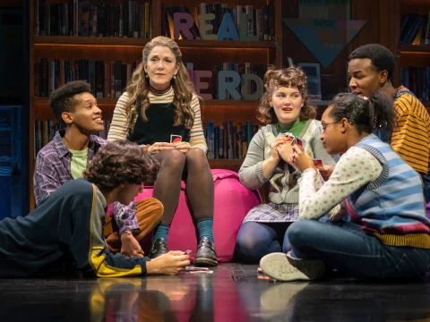 Group of young people and one adult sitting in a circle on a stage, playing with cards and engaging in conversation in front of a bookcase with the brightly colored letters "READ NERD" displayed.