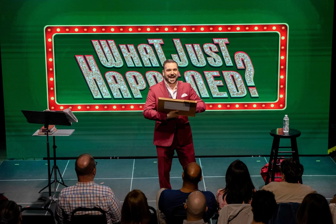 A man in a red suit stands on stage holding a box, with "What Just Happened?" displayed on a screen behind him. An audience watches from seated positions.