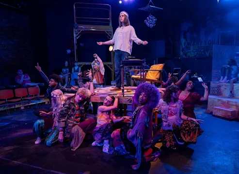 A group of actors in colorful costumes perform on stage, with one person standing on a table as others pose dramatically around them under blue and purple lighting.