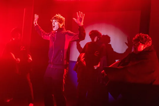 A person stands with raised hands under red lighting, surrounded by three others in dark clothing with stylized “Z” symbols.