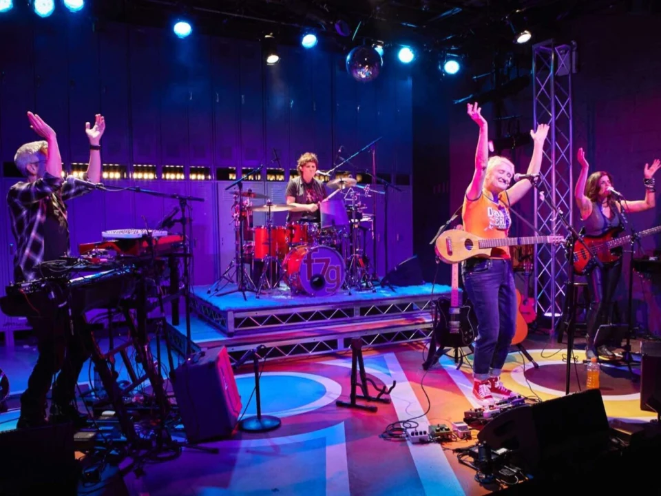 A band performs energetically on stage with drums, keyboards, and guitars under colorful lights.