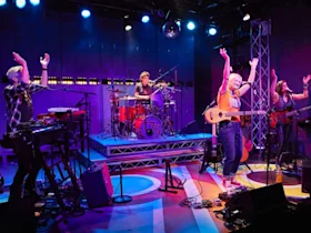 A band performs energetically on stage with drums, keyboards, and guitars under colorful lights.