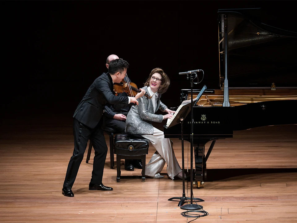 A woman plays a grand piano on stage, smiling at a man standing beside her. A violinist is seated behind her. Microphones are set up nearby.