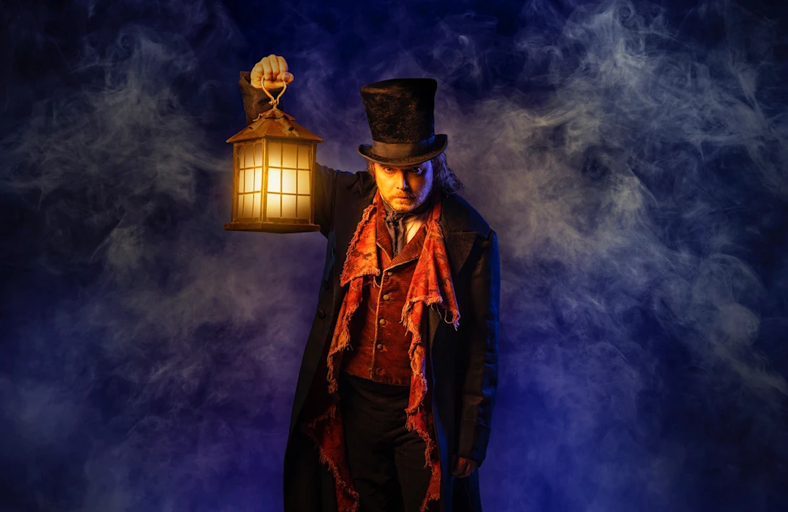 A man in Victorian-style clothing and a top hat holds a lit lantern, standing against a smoky, dark blue background.