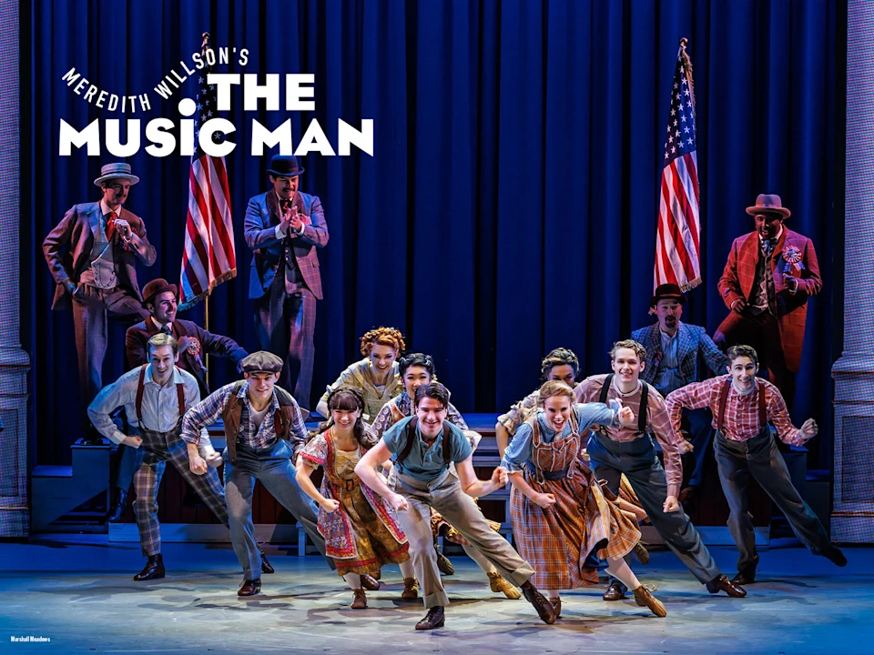 A group of actors in period costumes perform energetically on stage with American flags and blue curtains in the background. “The Music Man” is displayed in large letters above them.