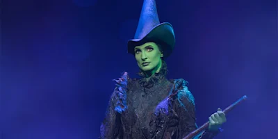 A person dressed as a witch with green face paint, a tall pointed hat, and holding a staff, stands against a blue-lit background.