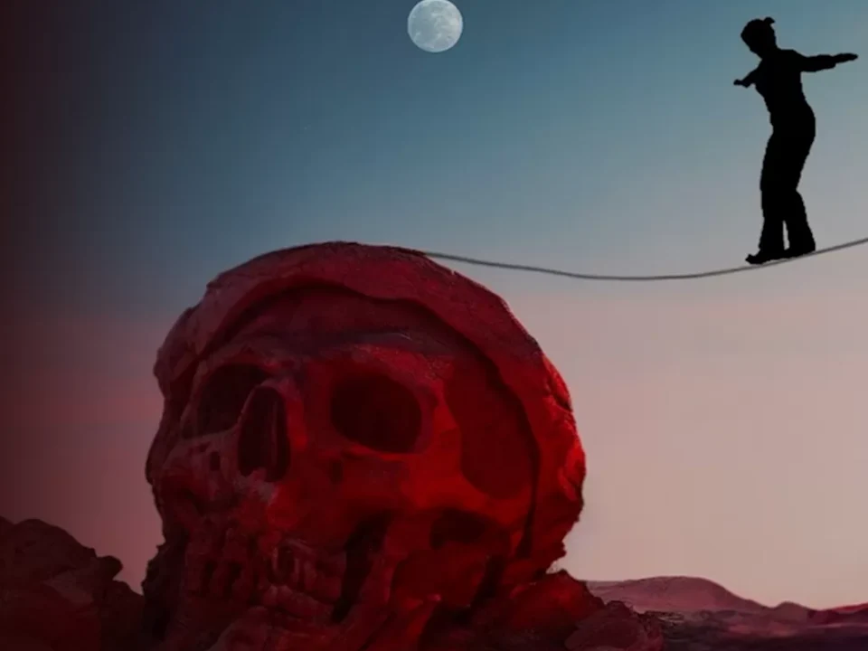 A person balances on a tightrope above a large red skull under a full moon.